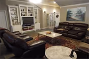A cozy living room with dark brown leather sofas arranged around a rectangular coffee table on a patterned rug. The room features built-in white shelves with decorative items and a flat-screen TV. There is a glass display cabinet and a large framed painting on the wall. The lighting is warm and the walls are painted in a neutral color.