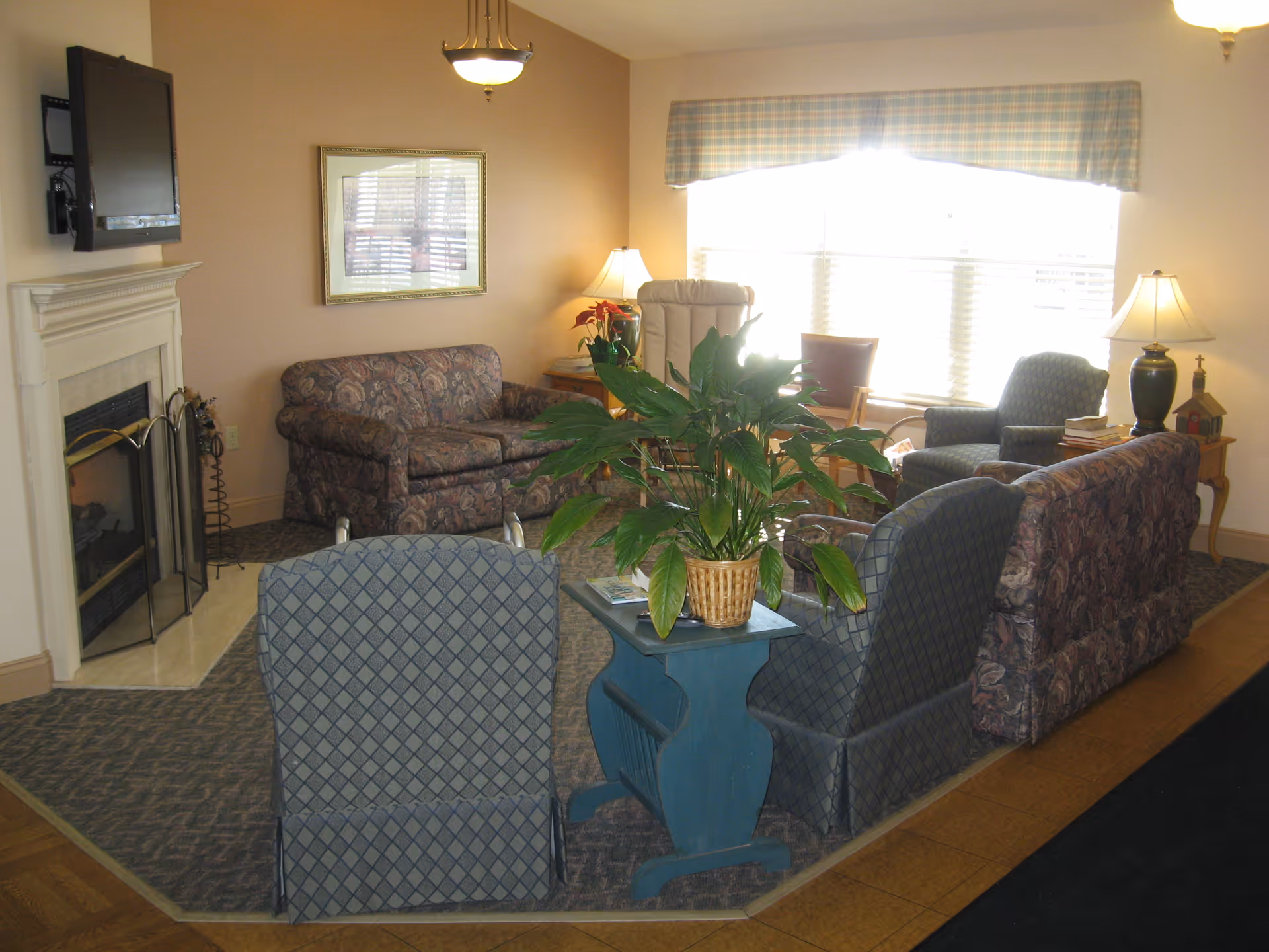 A cozy living room area in a senior living community featuring a floral-patterned sofa, two blue patterned armchairs, a beige recliner, and a wooden chair near a large window with blinds and a plaid valance. There is a fireplace with a mounted flat-screen TV above it, a blue side table with a potted plant, two table lamps, and framed artwork on the wall.