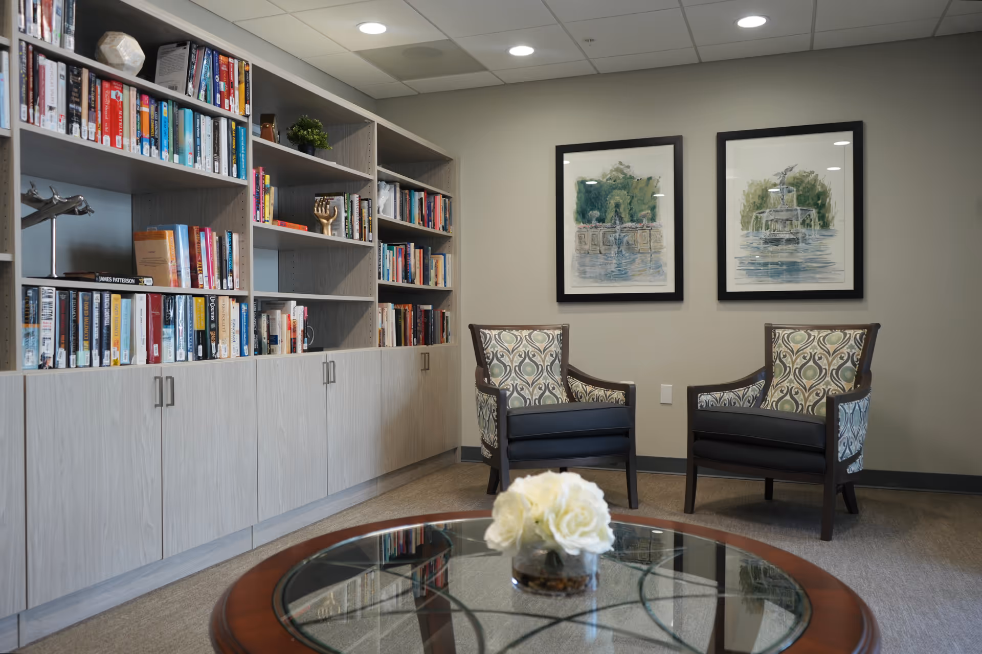 Seating area with built-in bookshelves, two patterned armchairs, framed artwork on the wall, and a glass-top table with flowers.
