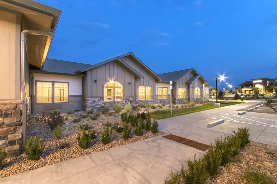Exterior view of a senior living facility building at dusk with lit windows, landscaped garden beds with small shrubs and rocks, a concrete sidewalk, and an adjacent parking lot with street lamps.