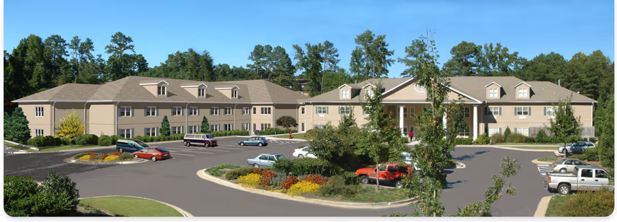 Front exterior of a large two-story beige assisted living building with a circular driveway, landscaped islands, and parked cars.