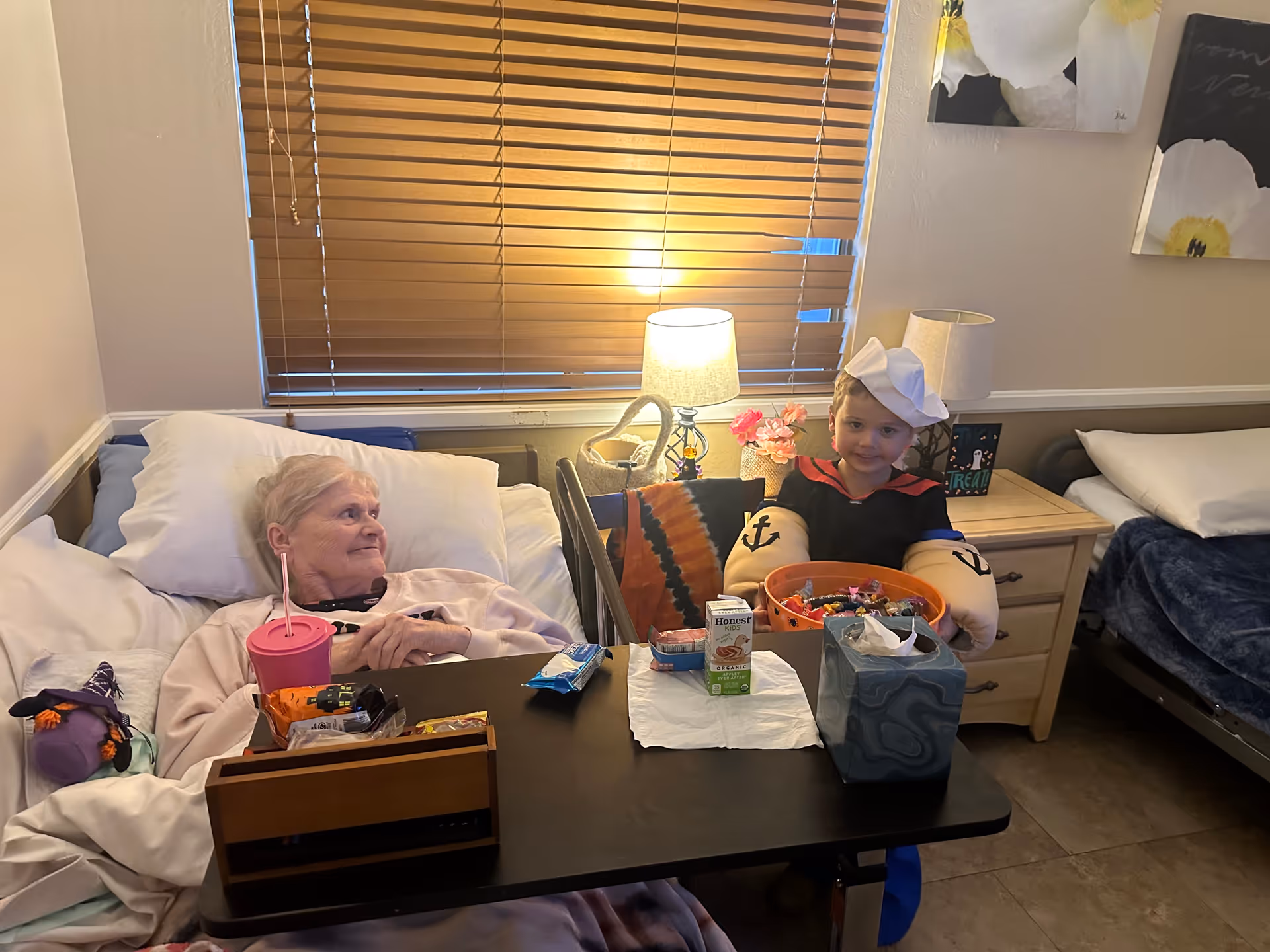 An elderly woman lying in a bed in a room with beige walls and a window with wooden blinds. She is looking at a young child dressed in a sailor costume sitting next to the bed. A table in front of the bed holds snacks, a pink cup with a straw, a juice box, and a tissue box. The room has another bed with white pillows and a blue blanket, a wooden nightstand with a lamp, flowers, and framed pictures. Two floral paintings hang on the wall.