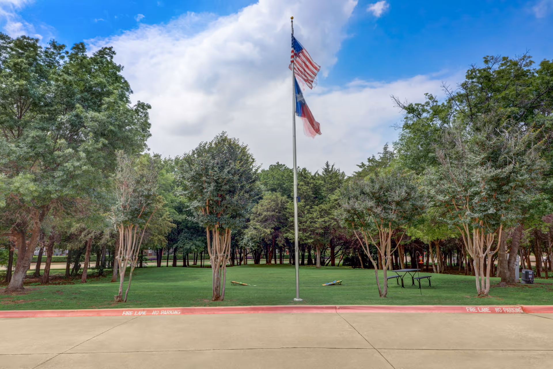 A grassy outdoor area with several trees surrounding a flagpole displaying the American and Texas flags. There are two cornhole boards on the grass and a picnic table to the right. The sky is partly cloudy with blue patches visible.