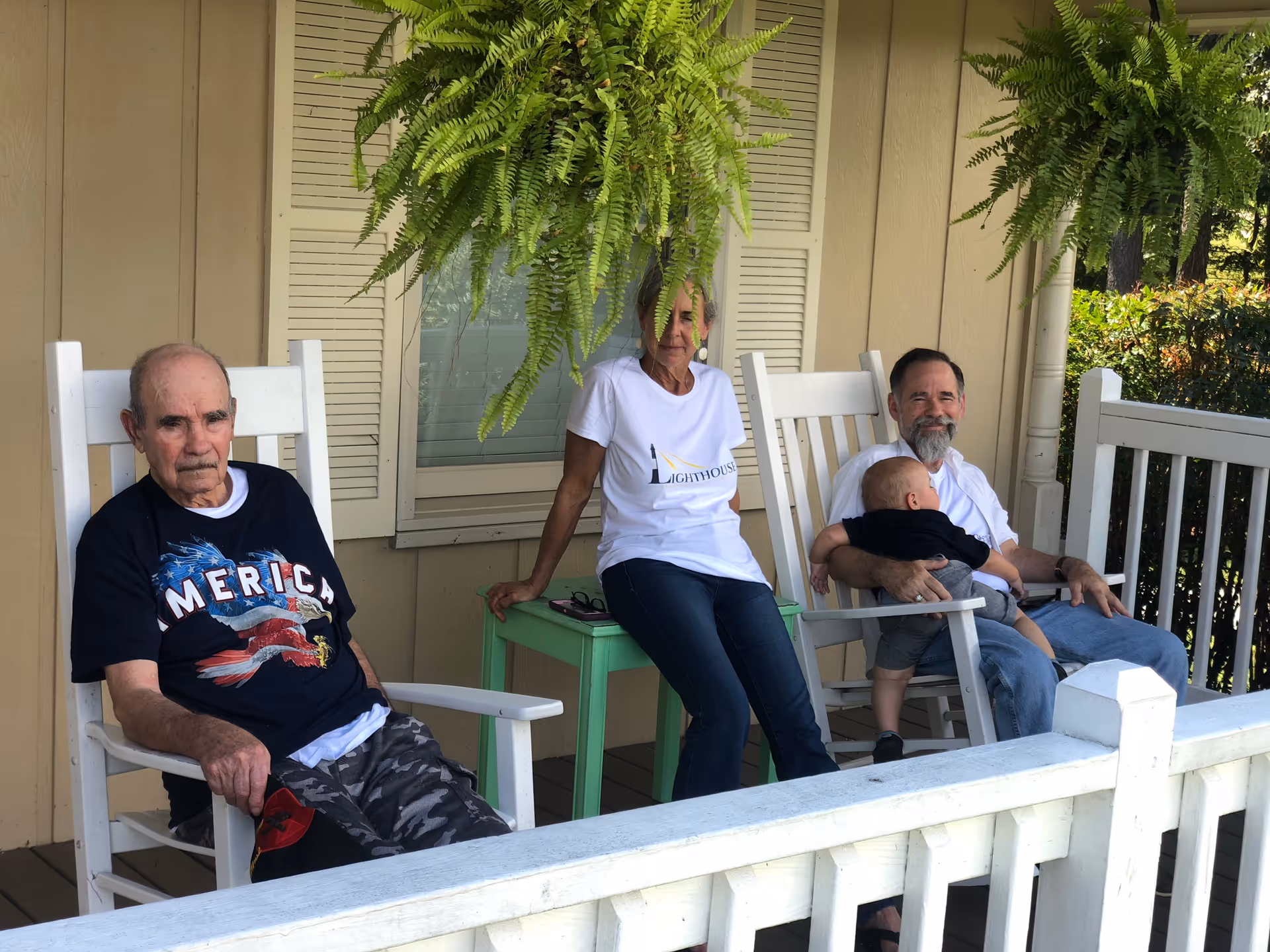Three adults and one baby sitting on white rocking chairs on a porch with beige siding and white railing. Two hanging green ferns are above them. One man is wearing a black 'America' t-shirt, a woman in a white 'Lighthouse' t-shirt is sitting in the middle, and another man is holding a baby on his lap.