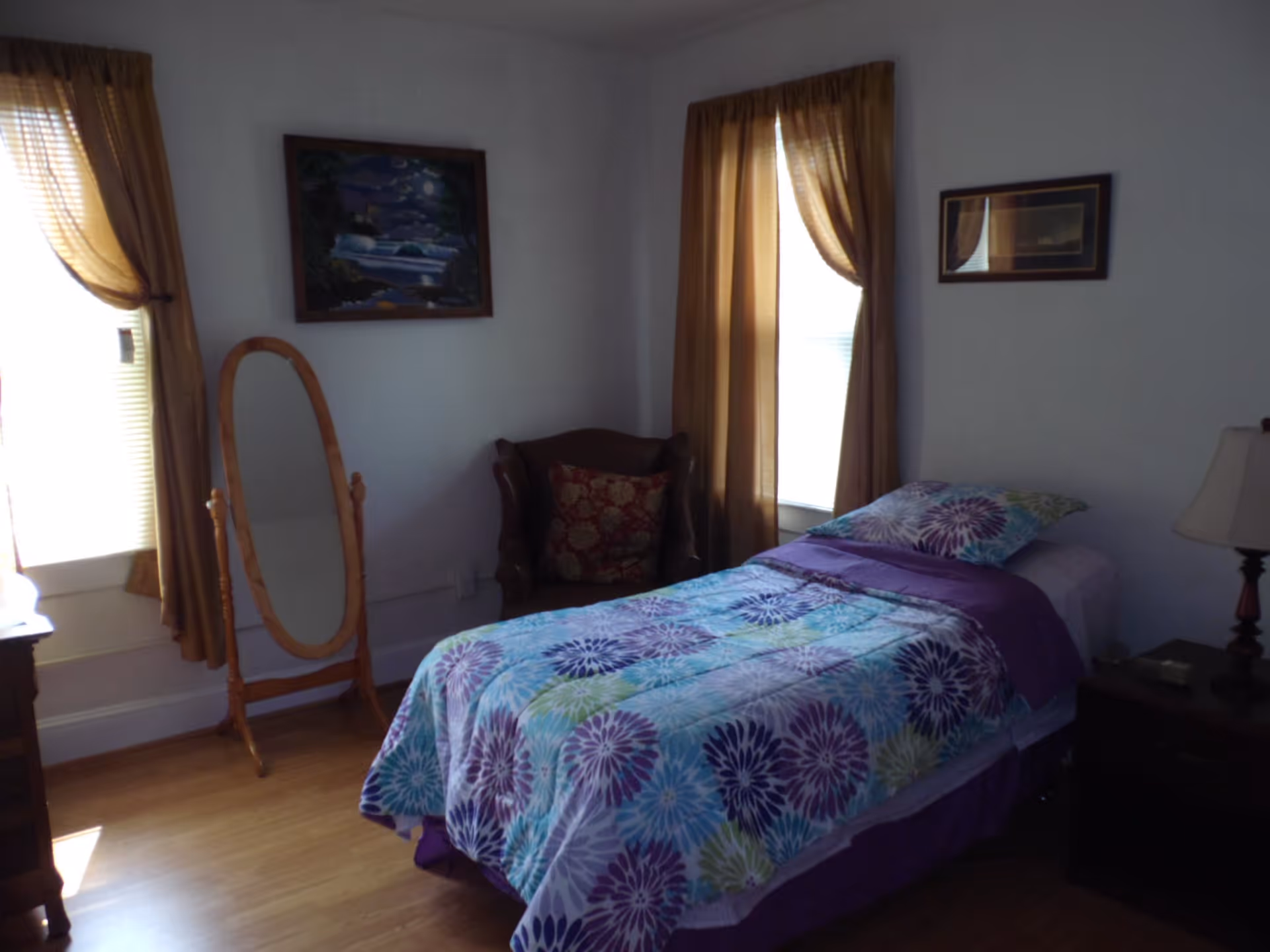 Small bedroom with a single bed dressed in a colorful floral comforter, a chair, floor mirror, and curtained windows.