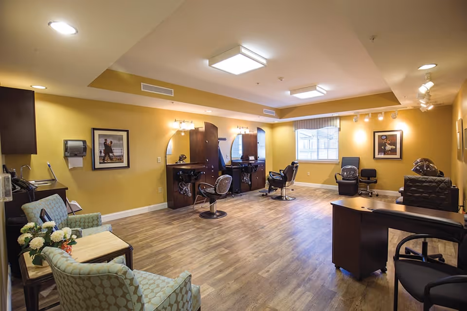 A spacious salon room with wooden flooring and yellow walls. The room features two salon chairs in front of mirrors with overhead lights, a sink area, and additional seating including armchairs and office chairs. There are framed pictures on the walls and a window with blinds letting in natural light.