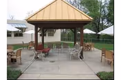 Outdoor patio area with a wooden gazebo structure in the center, surrounded by several chairs and tables on a concrete surface. The background shows a building and some trees.