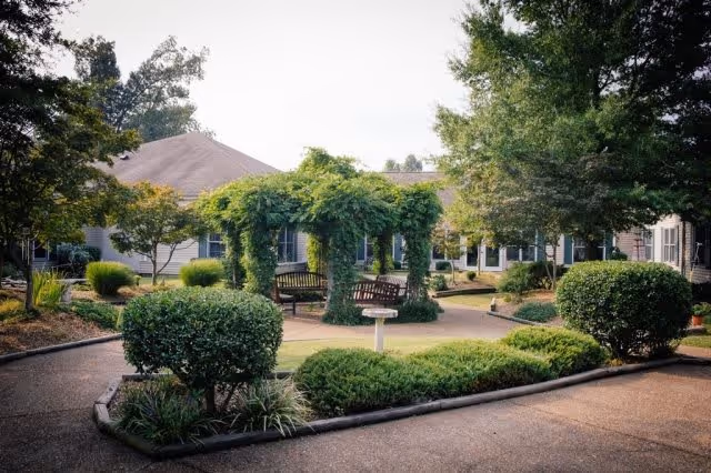 Outdoor garden area at Gaither Suites At West Park Assisted Living and Personal Care Home featuring a paved walkway, trimmed bushes, a wooden pergola covered with greenery, benches, and surrounding trees and shrubs.
