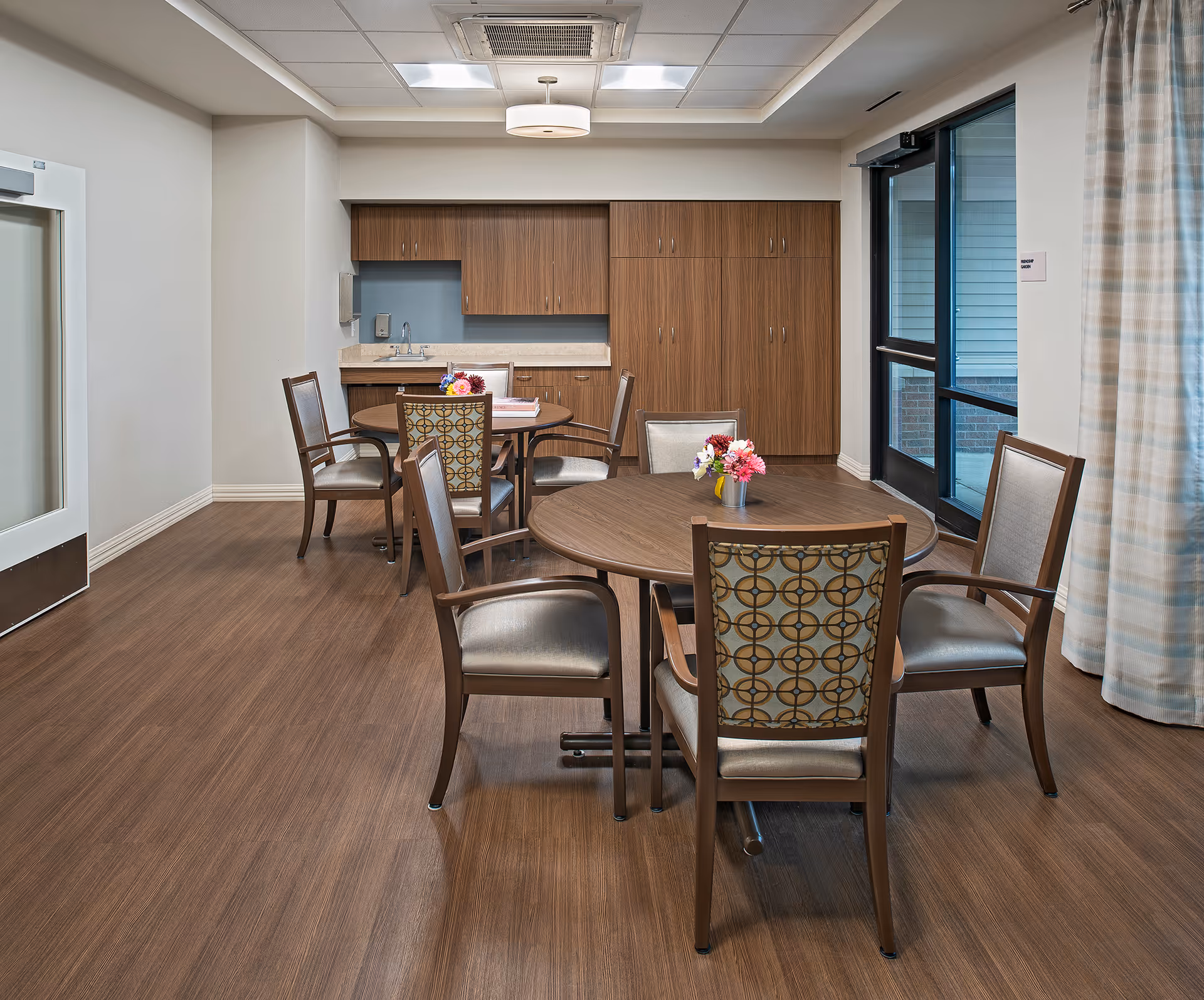A dining area in a senior living facility with two round wooden tables, each surrounded by four chairs with patterned backs and cushioned seats. There are small flower arrangements on each table. The room has wood flooring, a kitchenette with wooden cabinets and a sink, and a glass door leading outside. Light-colored walls and ceiling with recessed lighting complete the space.