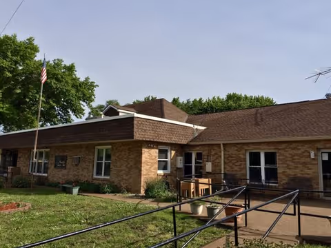 Single-story brick healthcare building with a wheelchair ramp, front lawn, and an American flag under a clear sky.