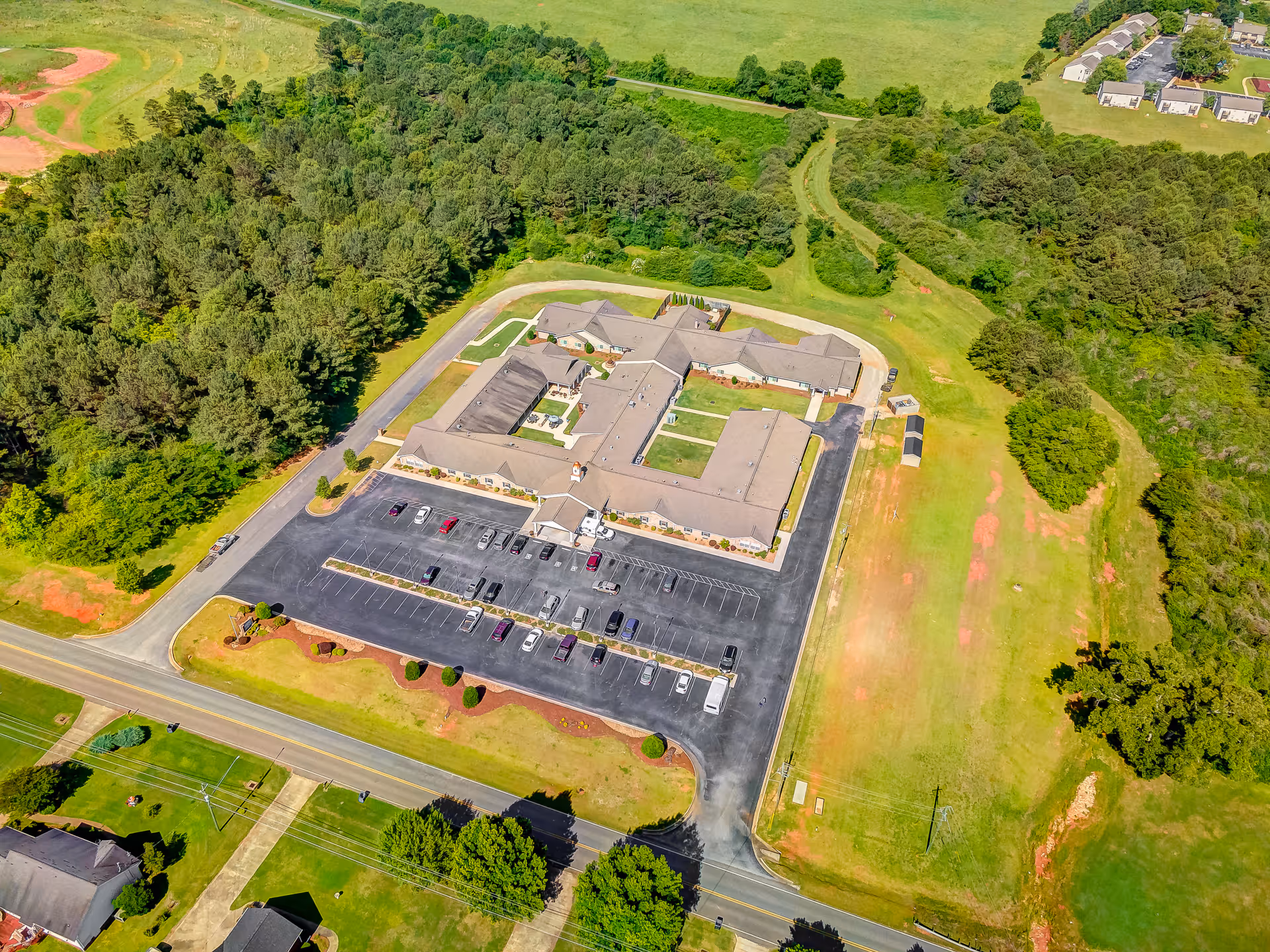 Aerial view of Legacy Village of Jacksonville facility showing a large single-story building with a U-shaped layout surrounded by parking lots, green lawns, and trees. The building is situated in a semi-rural area with open fields and some residential houses nearby.
