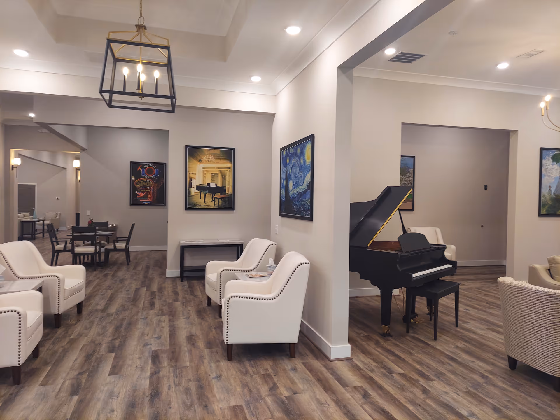 A spacious and well-lit common area with wood flooring, white cushioned armchairs, a black grand piano, and several framed artworks on the walls. There are tables and chairs in the background, and modern light fixtures hang from the ceiling.