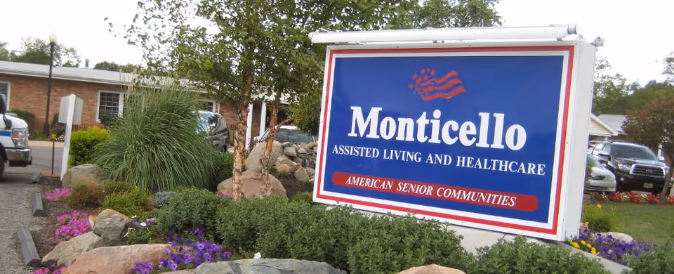 Large blue Monticello Assisted Living and Healthcare sign in a landscaped front area with flowers and parked cars.