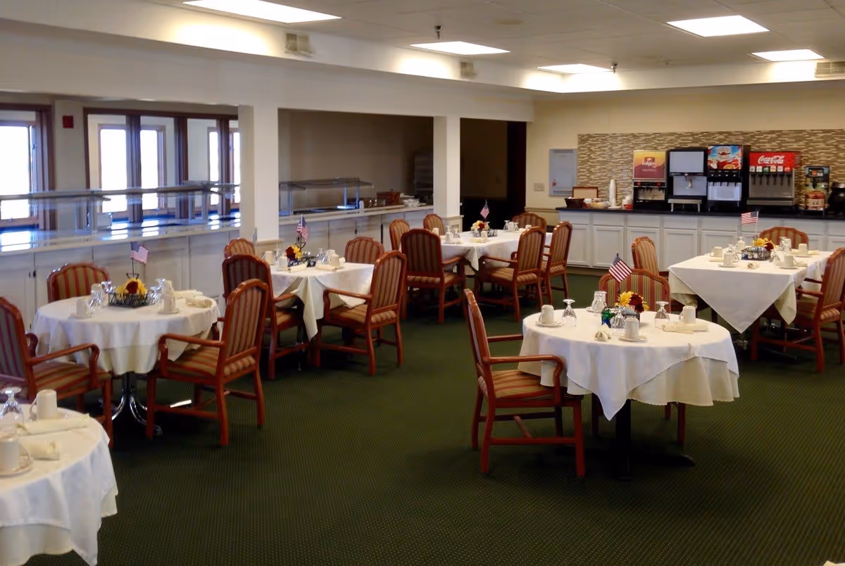 Dining room with round tables covered with white tablecloths, each set with cups, glasses, napkins, and small flower centerpieces with American flags. Chairs with striped upholstery surround the tables. In the background, there is a beverage station with soda dispensers and a coffee machine.