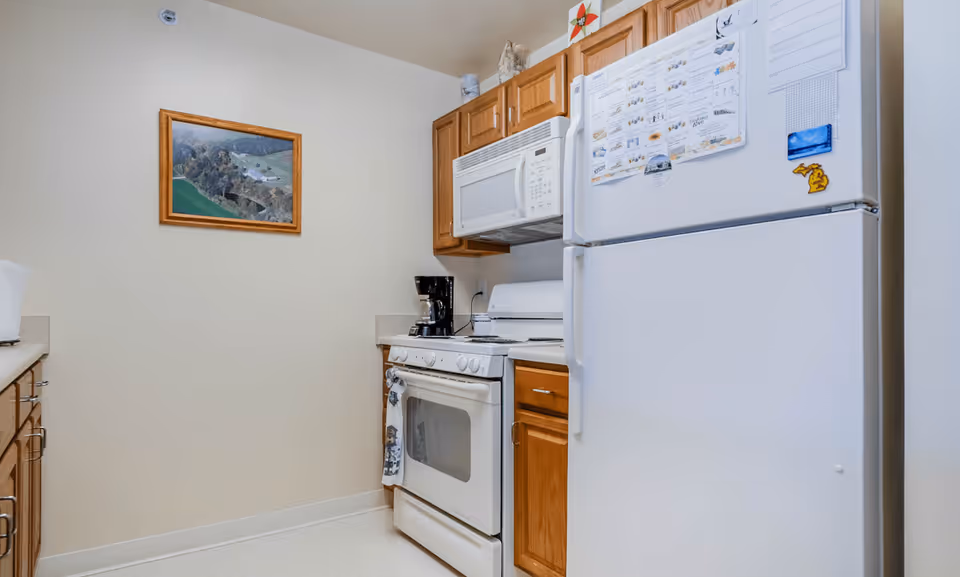 A small kitchen with wooden cabinets, a white refrigerator with magnets and papers attached, a white microwave above a white stove, a coffee maker on the counter, and a framed landscape picture on the wall.