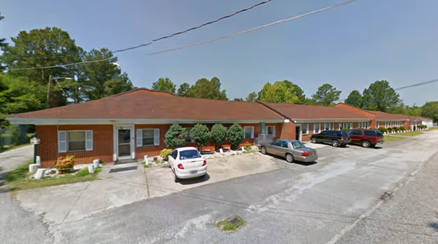 Exterior view of a single-story brick building with a red roof, several windows, and a few parked cars in front. The building is surrounded by trees and has a paved parking area.