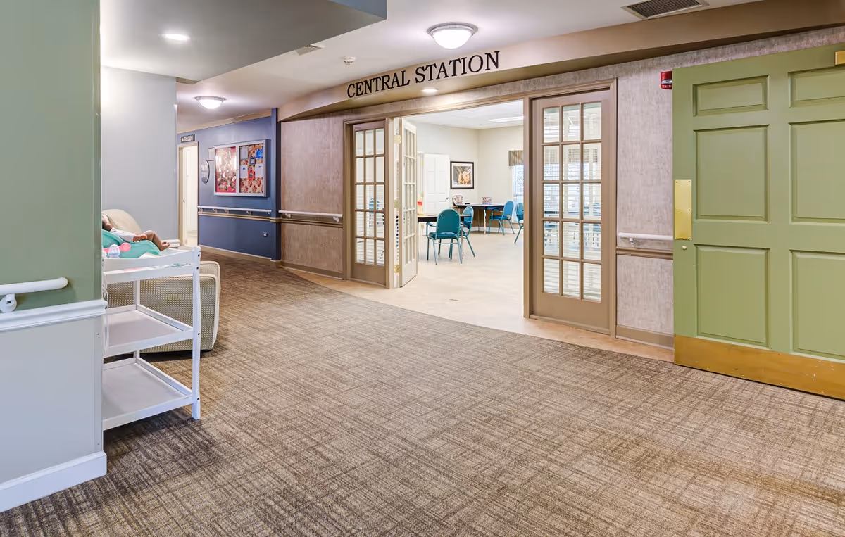 Carpeted interior hallway in a senior memory care facility leading to an open room labeled 'Central Station' with tables and chairs.