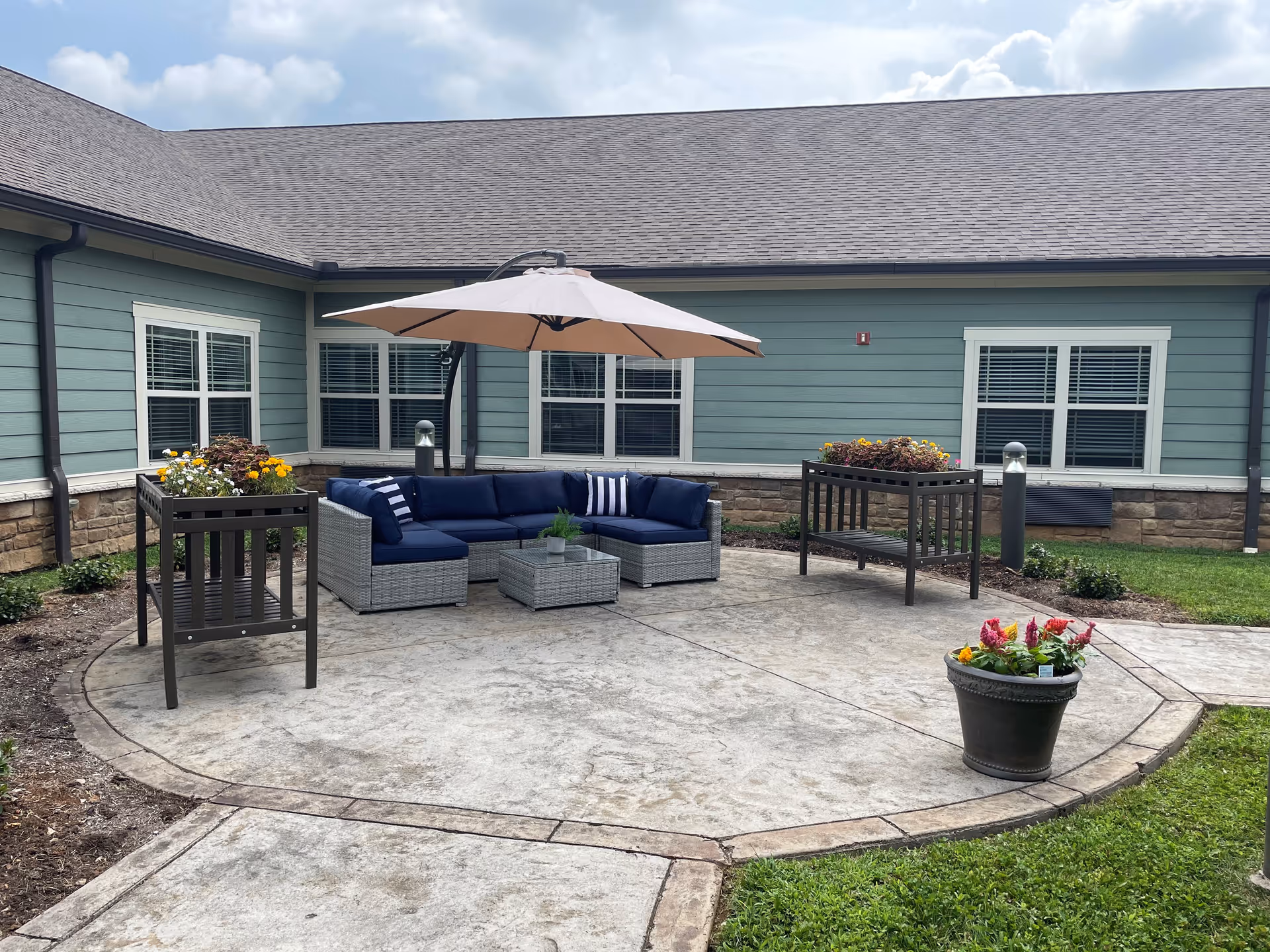 Outdoor patio area with a gray wicker sectional sofa with navy blue cushions and striped pillows under a large beige umbrella. There are two planter stands with flowers and a flower pot on the concrete patio. The patio is adjacent to a building with green siding, stone trim, and multiple windows with white blinds. The sky is partly cloudy.