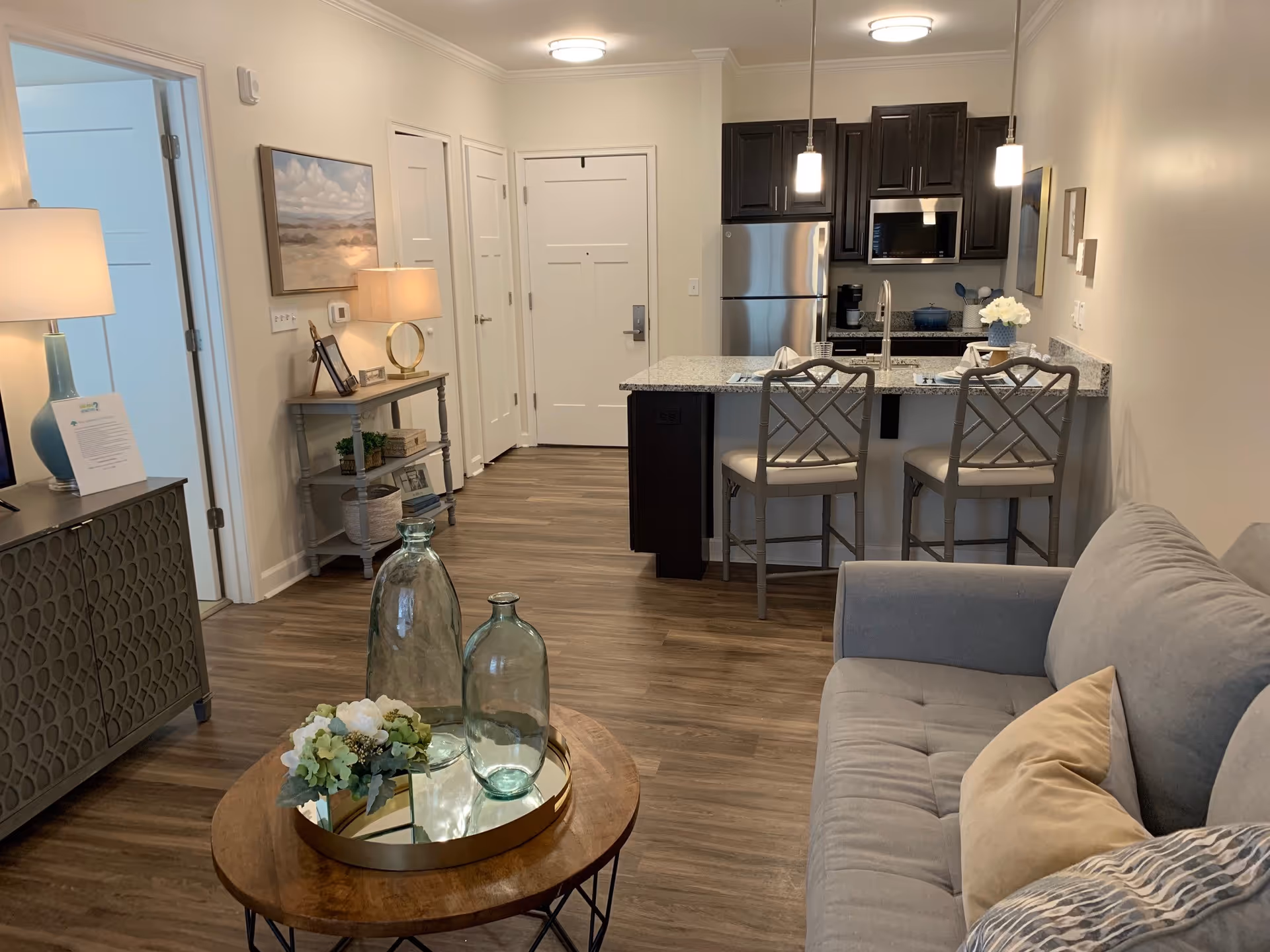 Open-plan living room and kitchen with a gray sofa, round coffee table with glass vases, and a granite-topped breakfast bar with two stools.