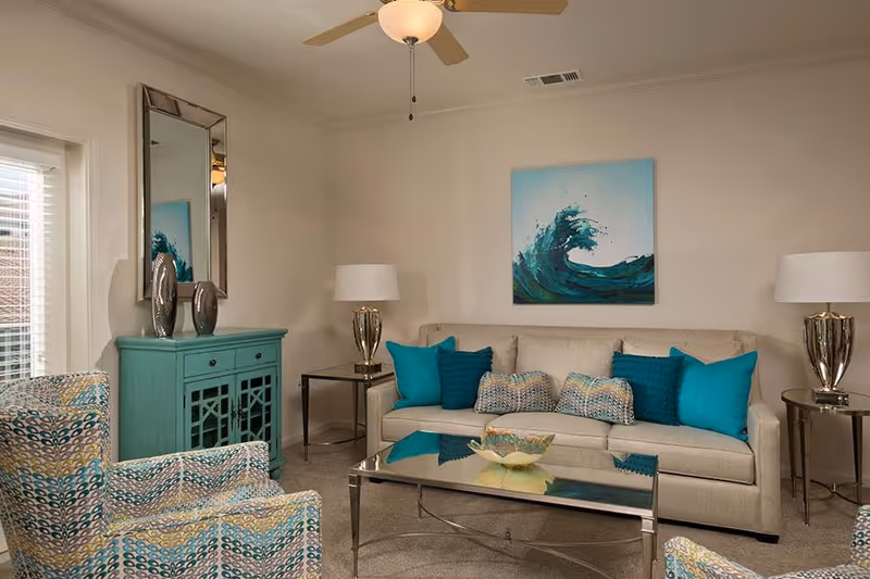 A cozy living room with a beige sofa adorned with teal and patterned throw pillows, two patterned armchairs, a glass coffee table with a decorative bowl, a teal cabinet with two silver vases, a large mirror above the cabinet, two side tables with matching lamps, and a ceiling fan. A painting of a wave hangs on the wall behind the sofa.