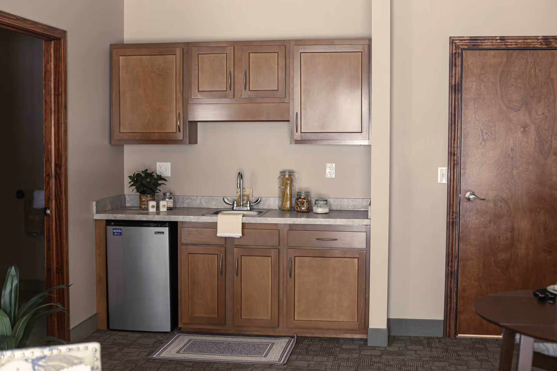 A small kitchenette area with wooden cabinets, a countertop with a sink, a small stainless steel refrigerator, and some jars and a plant on the counter. There is a wooden door to the right and a doorway to the left leading to another room.