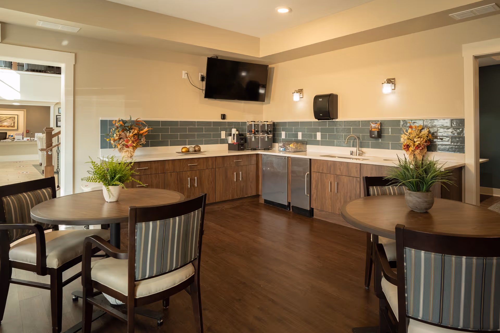 Small dining area with round tables and chairs facing a kitchenette with wood cabinets, green tile backsplash, sink, and a wall-mounted TV.
