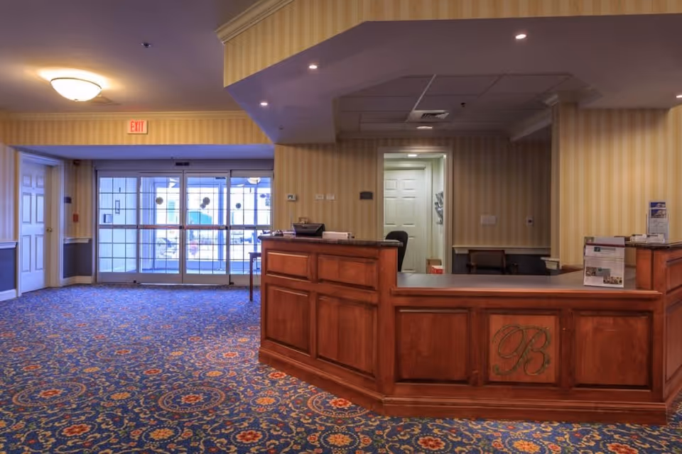 Reception area with a wooden front desk featuring a decorative 'B' emblem, patterned blue carpet with floral designs, yellow striped wallpaper, and a glass entrance door with an exit sign above it.
