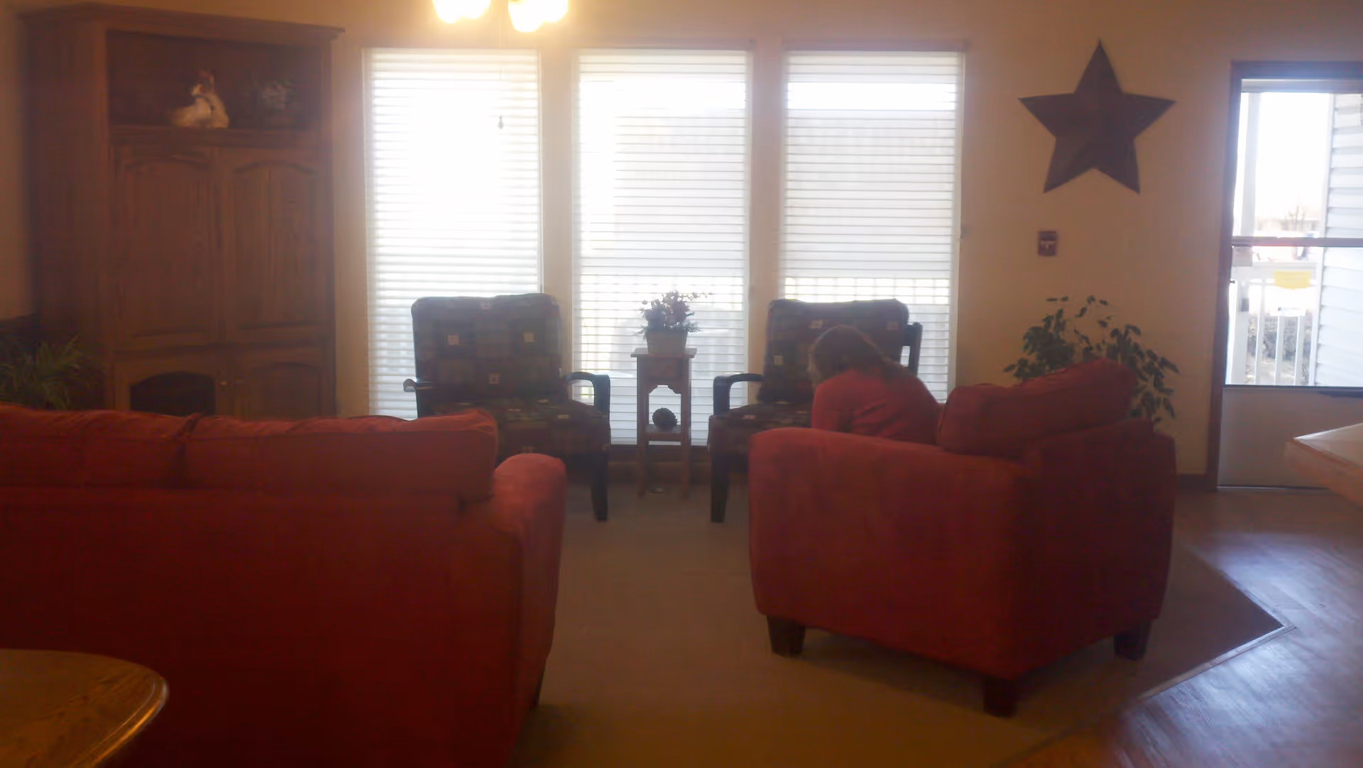 A cozy living room area with two red sofas facing two patterned armchairs separated by a small wooden table with a flower pot. There is a wooden cabinet on the left and a large star decoration on the wall to the right near a door with a window. A person in a red shirt is sitting on one of the red sofas, leaning forward.