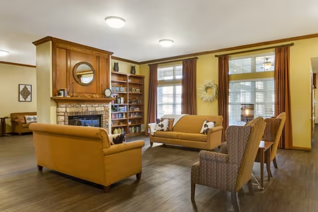 A cozy living room area with two mustard yellow sofas facing each other, a wooden fireplace with a round mirror above it, a bookshelf filled with books, and two patterned armchairs with a small table between them. Large windows with brown curtains allow natural light to fill the room.