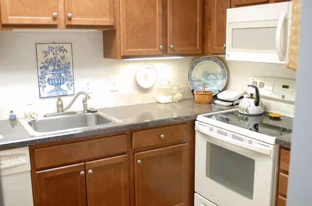 A kitchen area with wooden cabinets, a stainless steel sink, a white electric stove with a kettle on top, a white microwave above the stove, and decorative plates on the countertop and wall.