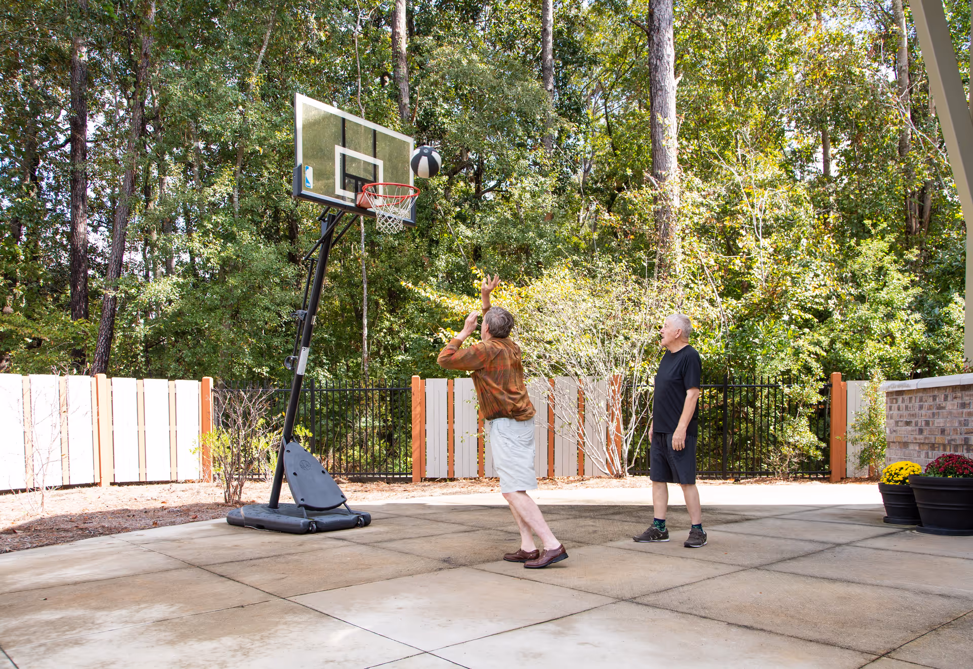 Two elderly men playing basketball outdoors on a concrete court surrounded by trees and a white fence. One man is shooting the basketball towards the hoop while the other watches.