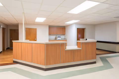 Reception desk area inside a healthcare or rehab facility with wood-paneled counters, white ceiling tiles with fluorescent lighting, and doors leading to other rooms in the background.