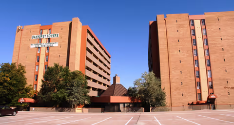 Exterior view of a retirement community named Oakhurst Towers, showing two large brick buildings with multiple floors and windows, a clear blue sky, some trees, and an empty parking lot in the foreground.