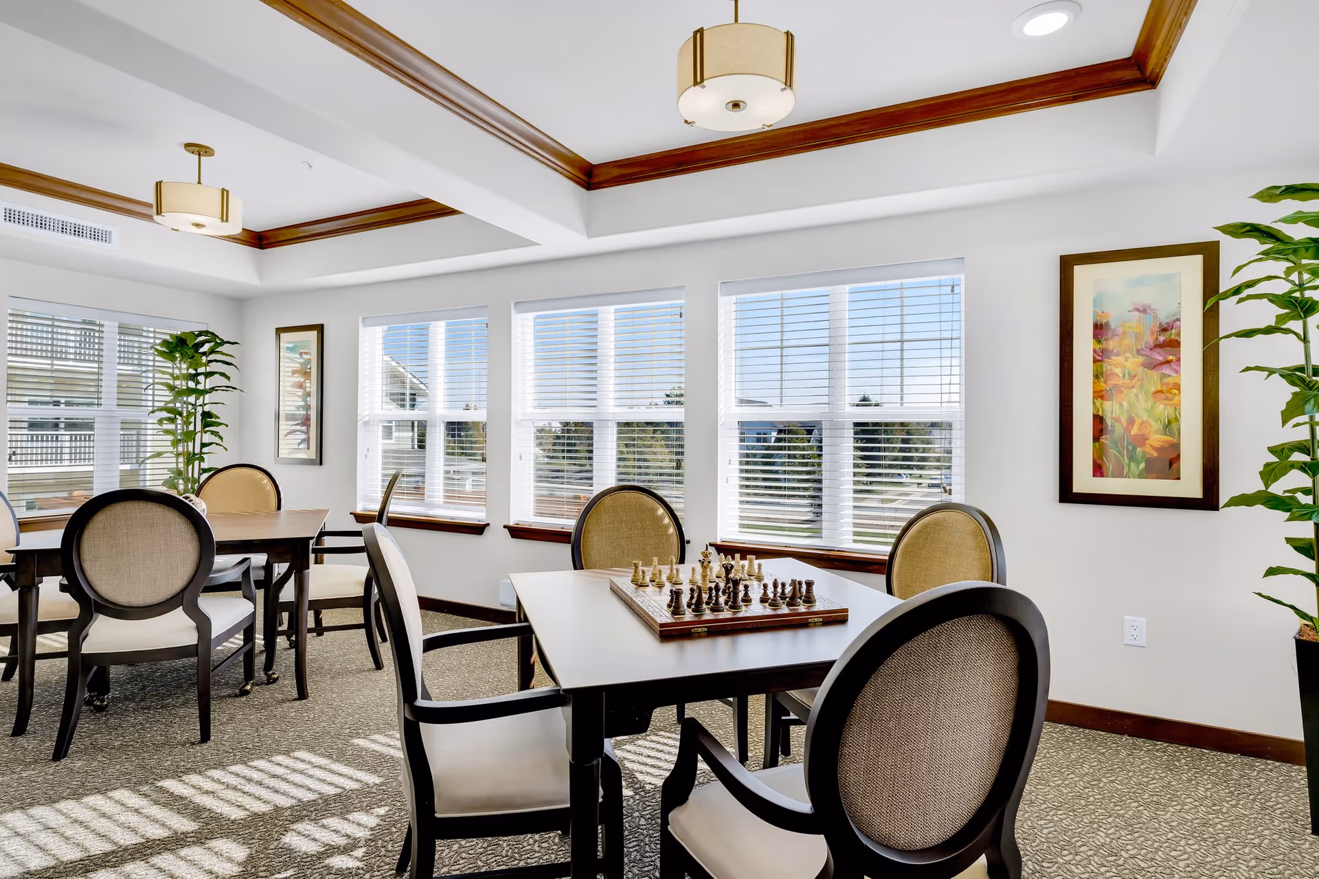 A bright and airy room with multiple tables and chairs arranged for social or recreational activities. One table has a chessboard set up for a game. The room features large windows with white blinds, allowing natural light to fill the space. There are two framed floral paintings on the walls and tall green plants in black pots in the corners. The ceiling has recessed lighting with wooden trim accents.