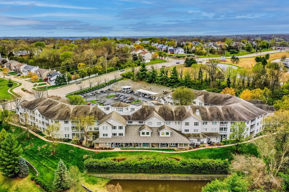 Aerial view of a large senior living facility named Solstice Senior Living at Fenton, surrounded by green lawns, trees, and a small pond in the foreground. The building has multiple wings with a parking lot and nearby residential neighborhood visible in the background under a partly cloudy sky.