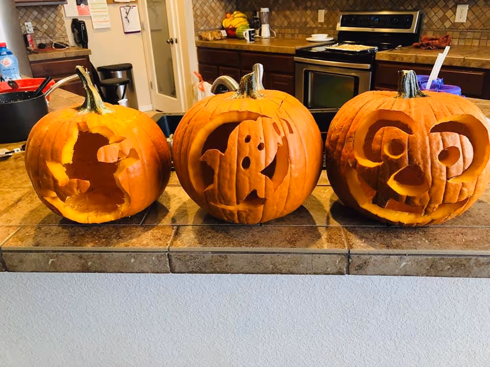 Three carved pumpkins displayed on a kitchen counter. The pumpkin on the left has a partially carved design, the middle pumpkin features a ghost carving, and the pumpkin on the right has a face with large eyes and a nose. The background shows a kitchen with a stove, cabinets, and various kitchen items.