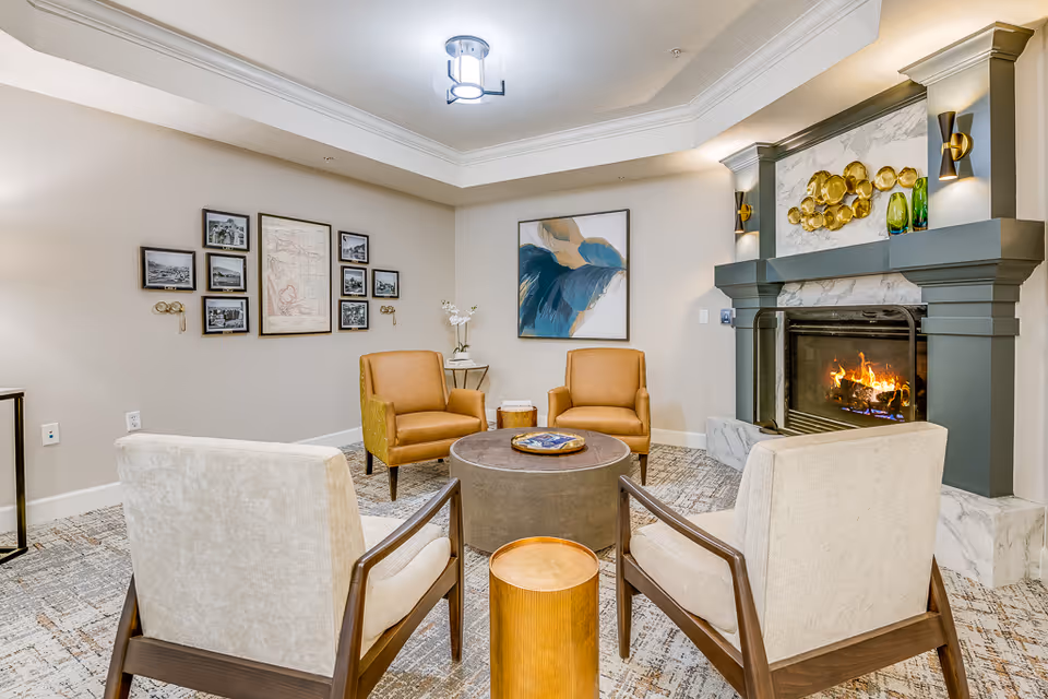 A cozy living room area with a lit fireplace, two tan armchairs, two beige armchairs, a round coffee table, and a small wooden side table. The walls are decorated with framed artwork and photographs. The room has a neutral color palette with soft lighting.
