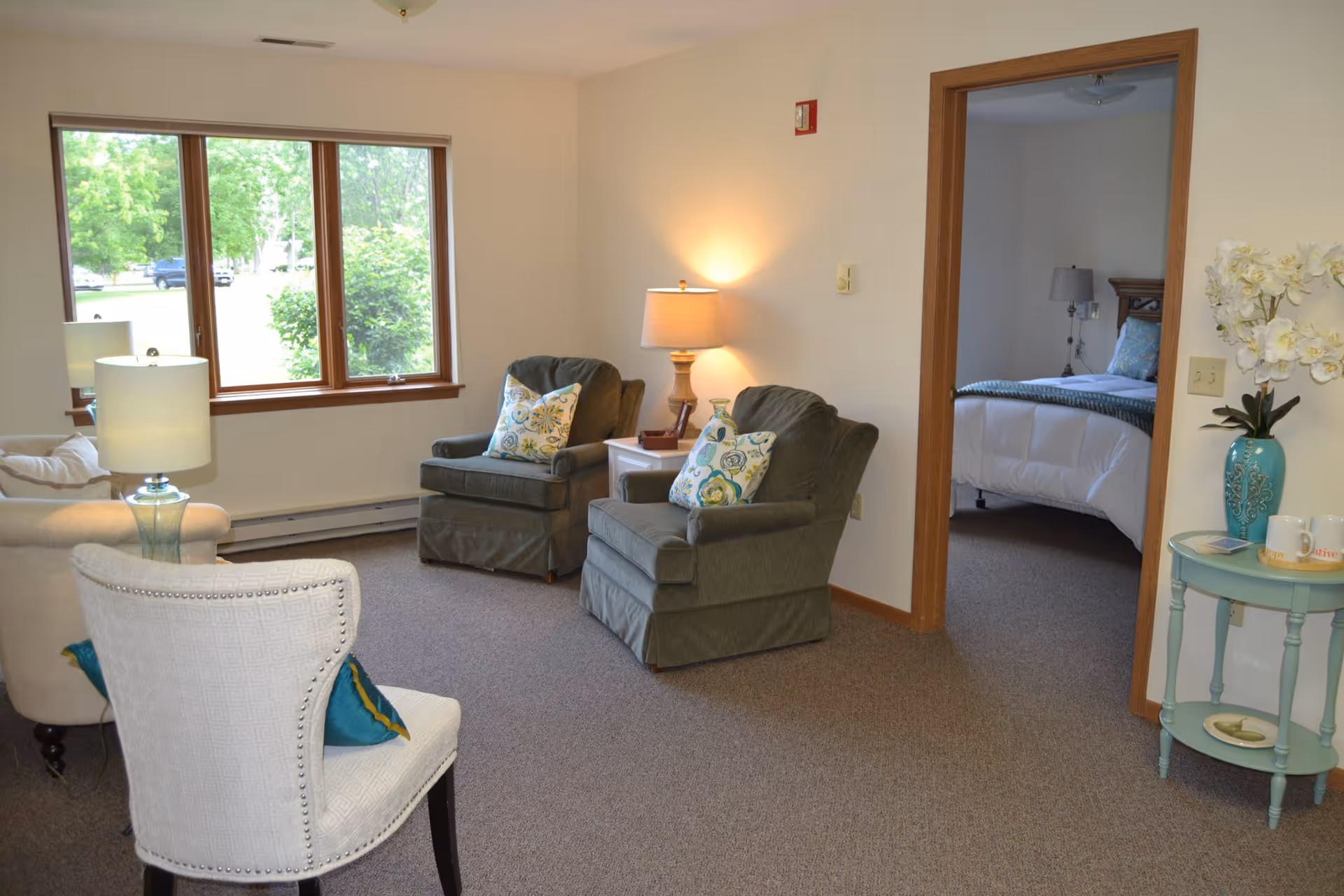 A cozy living room area with two green armchairs, a white armchair, and a beige sofa. There are two lamps providing warm light, a window showing greenery outside, and a small round table with a vase of white flowers and two mugs. An open doorway reveals a bedroom with a bed and a nightstand with a lamp.