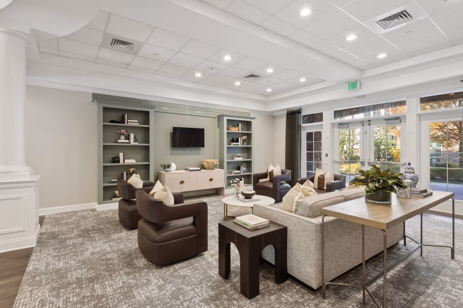A bright and spacious senior living facility lounge area with comfortable seating including a sofa and four armchairs arranged around a round coffee table. The room features built-in shelves with decorative items, a wall-mounted flat screen TV, large windows and glass doors letting in natural light, and a table with a plant and decorative vases behind the sofa.