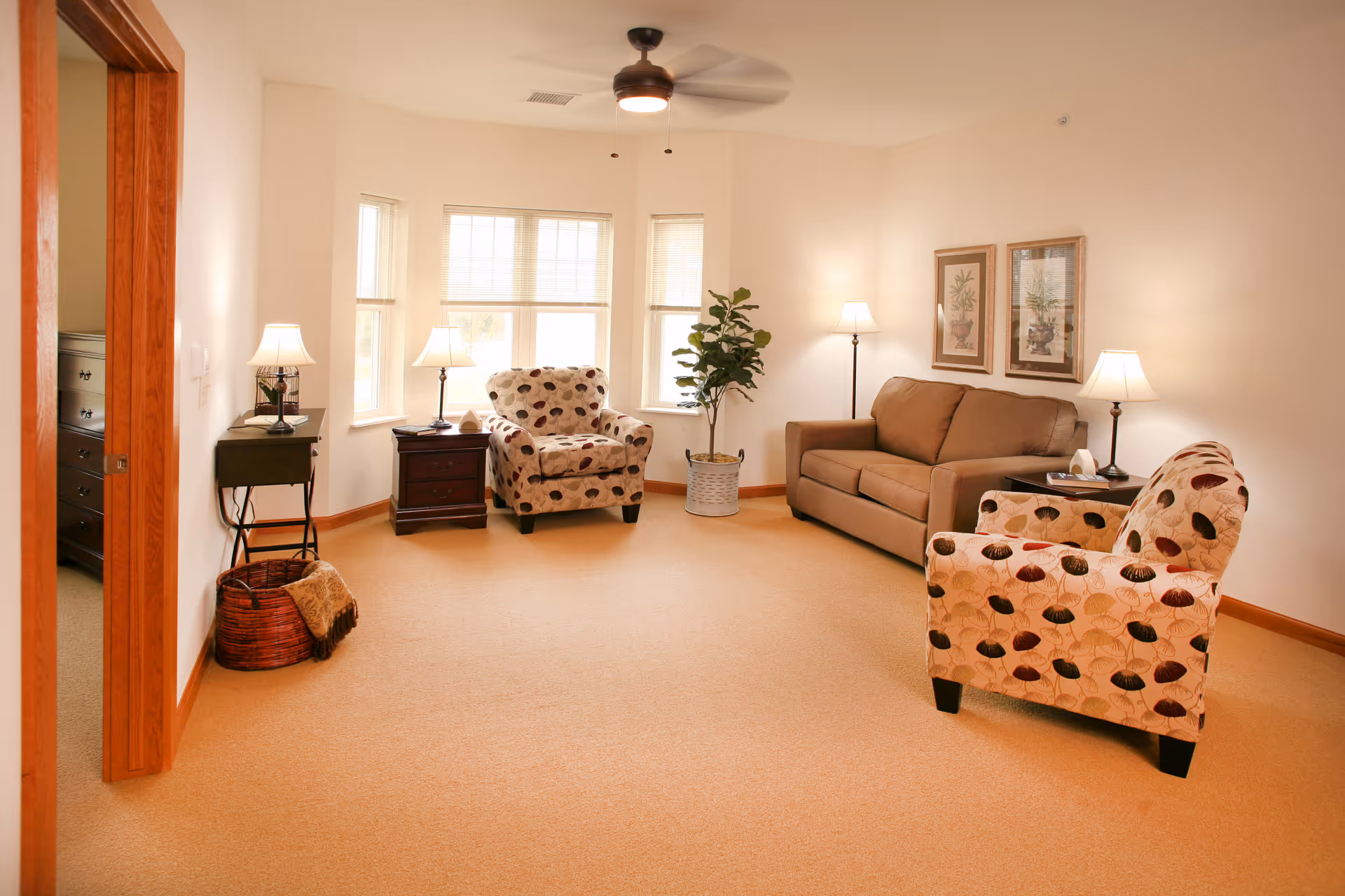 Well-lit living room with a sofa, two patterned armchairs, side tables and lamps, and a potted plant by bay windows.