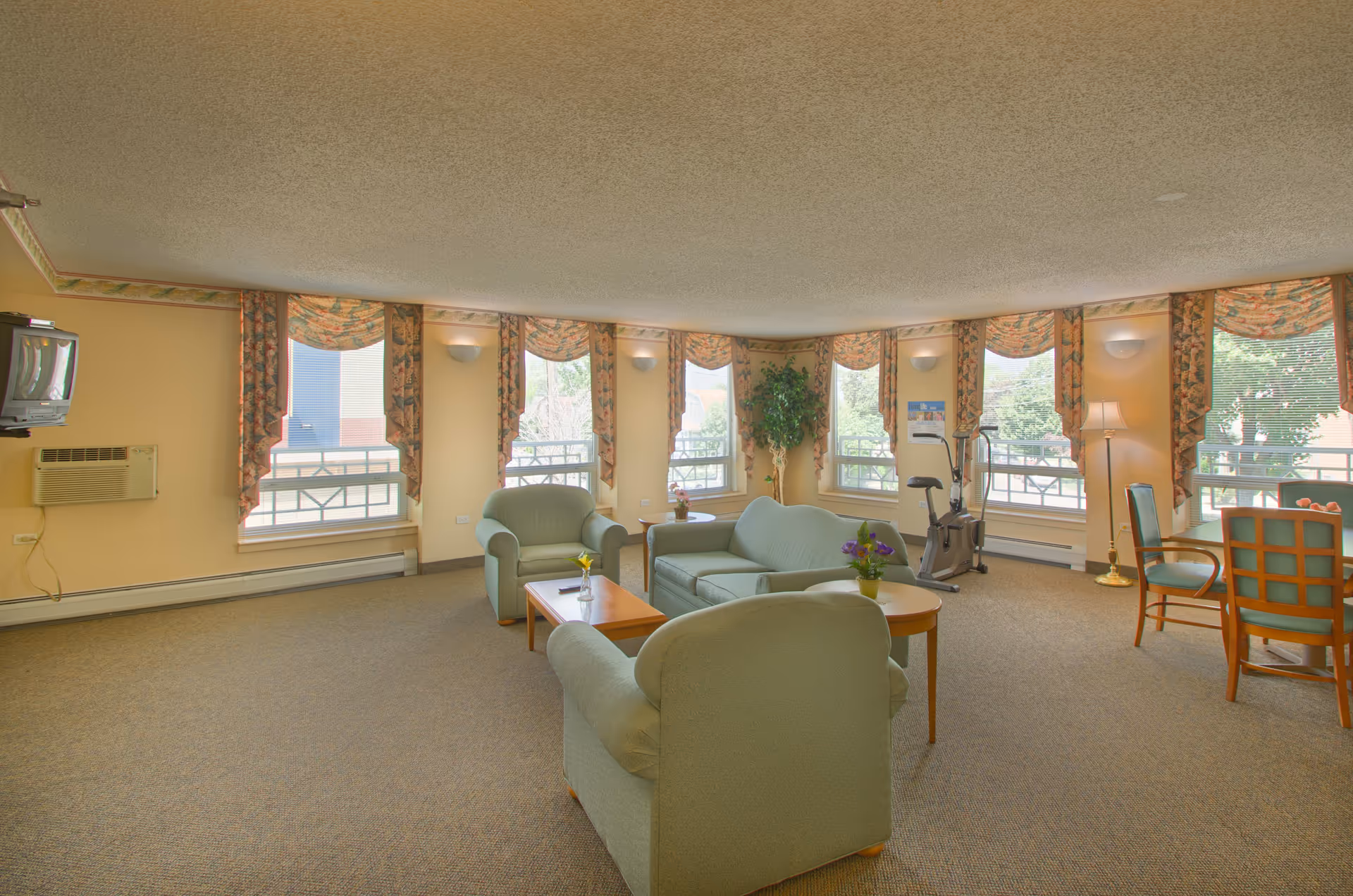 A spacious senior living common area with multiple large windows covered by floral valances, light green upholstered armchairs and a sofa arranged around wooden coffee tables with small flower vases. There is an exercise bike near the windows and a dining table with chairs in the corner. The room has beige walls, carpeted floor, a wall-mounted TV, and an air conditioning unit.