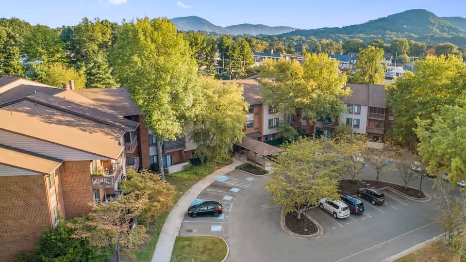 Aerial view of The Park Oak Grove showing multi-story brick residential buildings, trees, a parking lot with several cars, and hills in the background.