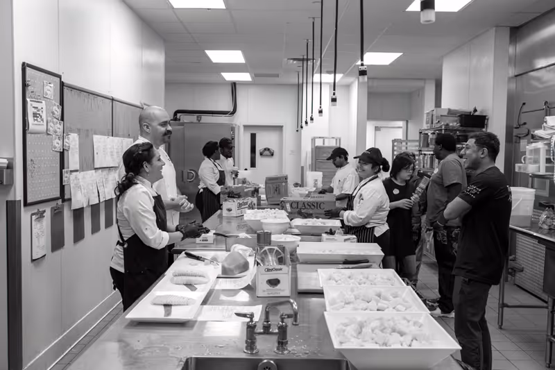 Black and white photo of a busy commercial kitchen with several people working and interacting. There are trays of food on the counter and kitchen equipment visible in the background.