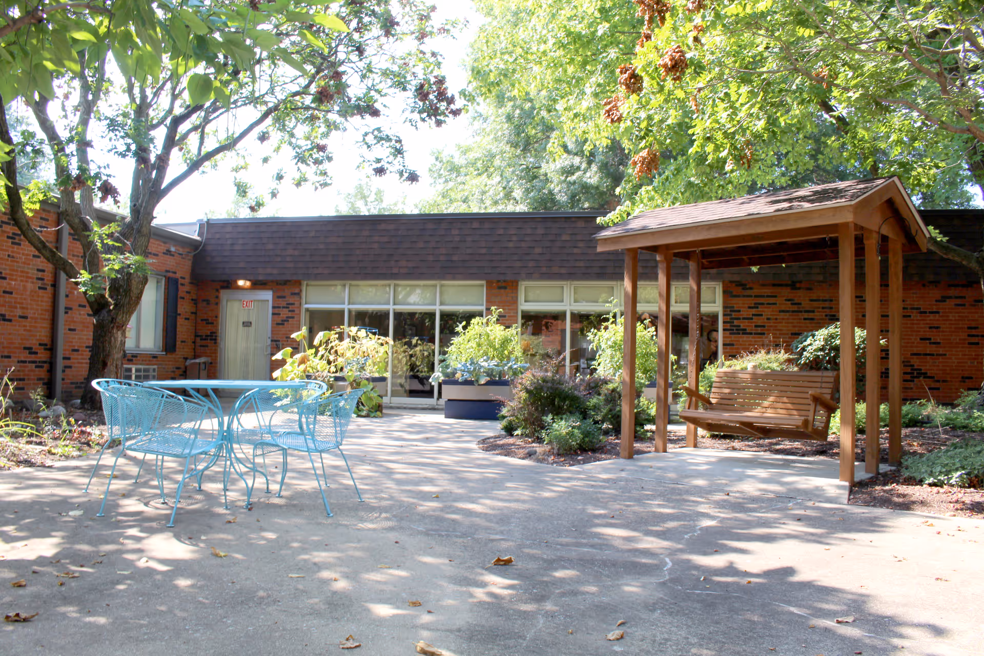 Outdoor patio area at Pana Health and Rehab Center featuring a blue metal table with four matching chairs, a wooden swing with a roof, surrounded by trees and plants, with a brick building in the background.