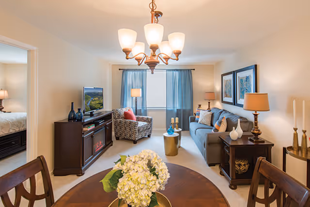A cozy living room in a senior living community featuring a gray sofa with decorative pillows, a patterned armchair, a wooden TV stand with a flat-screen TV, two side tables with lamps, and a round dining table with a floral centerpiece. Blue curtains frame a window in the background, and a bedroom is partially visible through an open doorway.