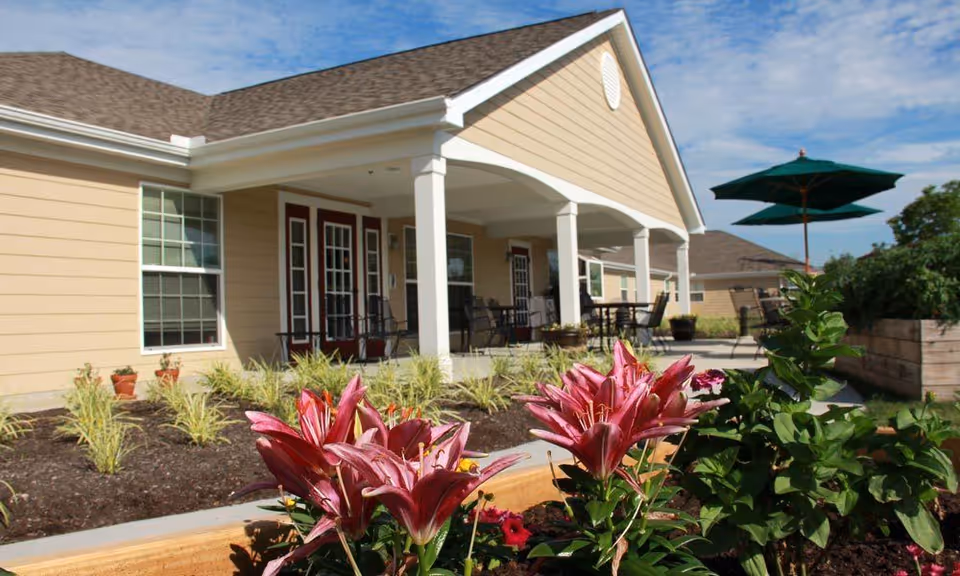 Exterior view of a single-story senior living building with a covered patio, outdoor seating, umbrella, and flowerbed with pink lilies in the foreground.