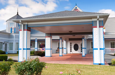 Covered porte-cochere and entrance of a white building with blue and red trim, landscaped lawn and flowering bushes in front.