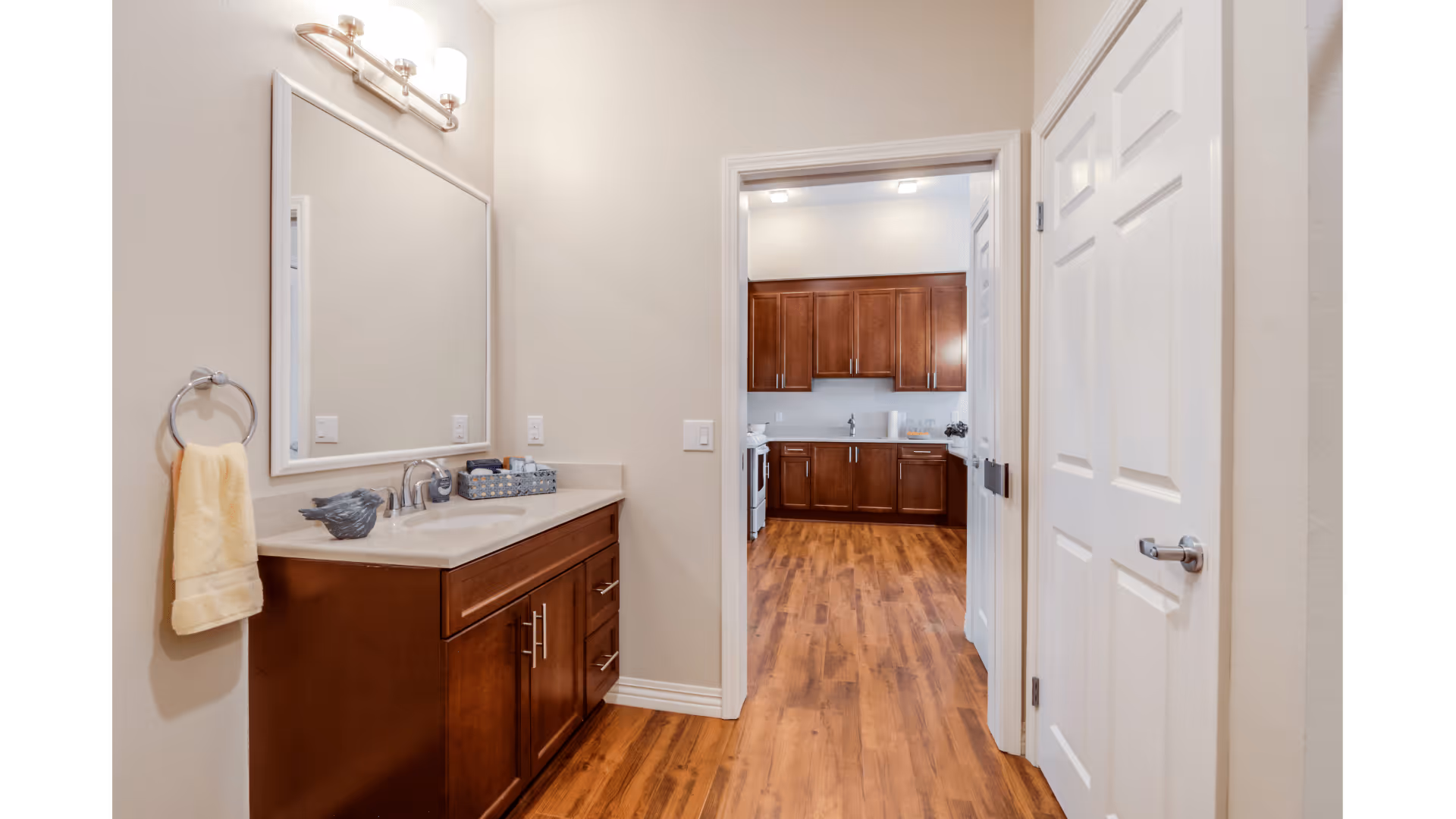 View of a bathroom vanity with a large mirror, countertop with a faucet, and a yellow towel hanging on a ring. The bathroom opens into a kitchen area with wooden cabinets and wood flooring.