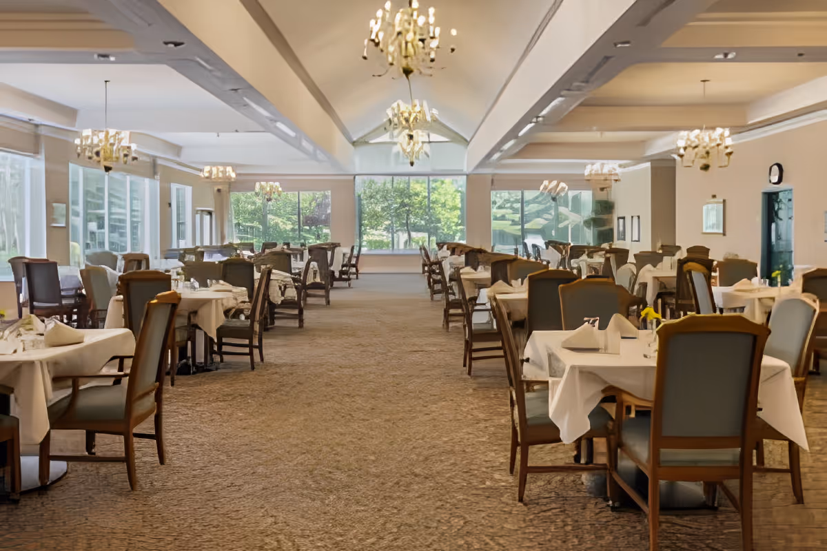 Spacious dining room with multiple tables covered in white tablecloths, each set with napkins and silverware. The room features large windows letting in natural light and chandeliers hanging from the ceiling. Chairs with wooden frames and cushioned seats surround the tables.