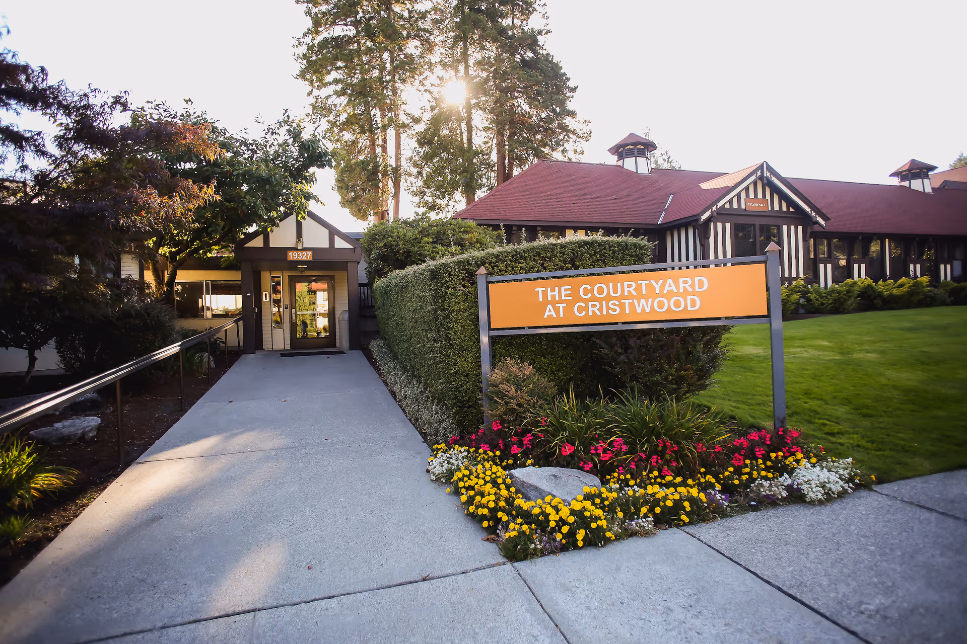 Walkway leading to the entrance of a care facility with a sign reading "The Courtyard at Cristwood" and flowerbeds in front.